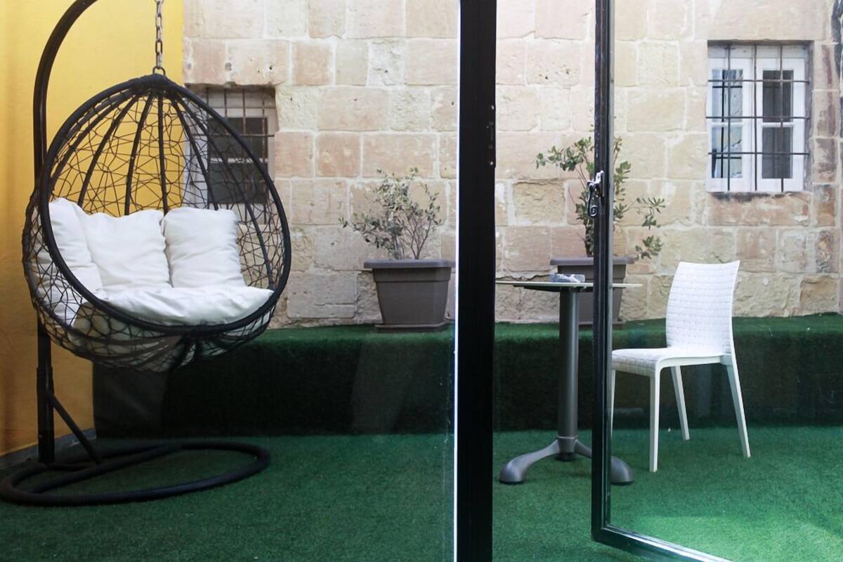 Una silla colgante con cojines, una silla blanca, una mesa y una planta en maceta en un patio con paredes de piedra y césped artificial.