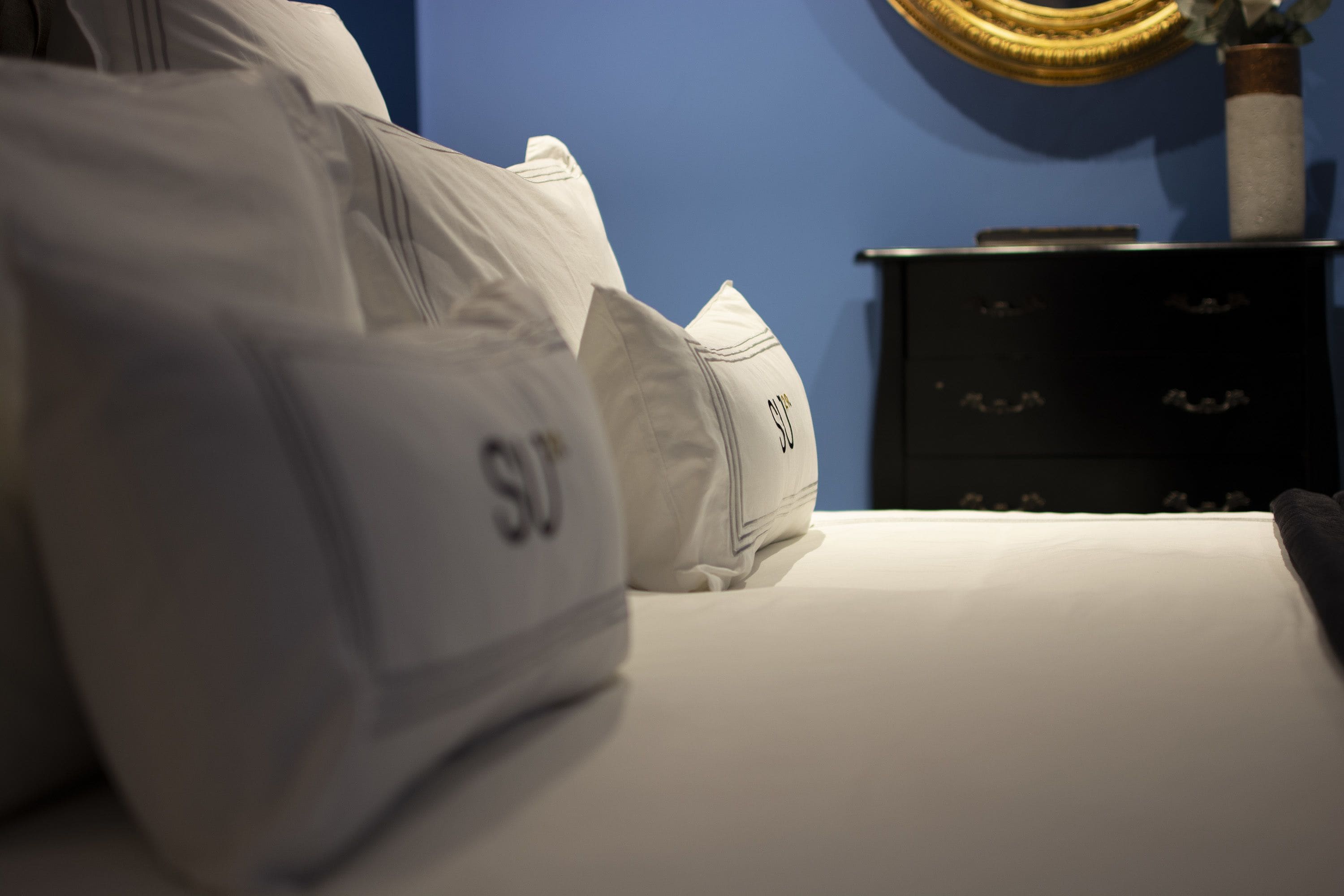 A bed with white bedding and pillows, a dark wooden dresser, and a blue wall with a decorative golden object.