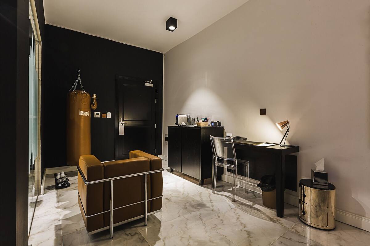 Una habitación de hotel moderna con un espacio para ejercitarse, un escritorio con computadora y un rincón de descanso con un saco de boxeo.