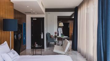 Una habitación de hotel moderna con una cama grande, un área de descanso con una silla y una mesita, un escritorio con silla, y un ventanal con cortinas.