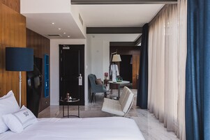 Una habitación de hotel moderna con una cama grande, un área de descanso con una silla y una mesita, un escritorio con silla, y un ventanal con cortinas.