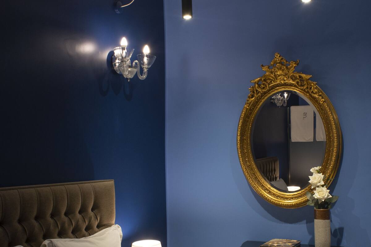Un dormitorio con cabecera capitonada, ropa de cama blanca, cómoda negra, espejo con marco dorado y aplique de pared.