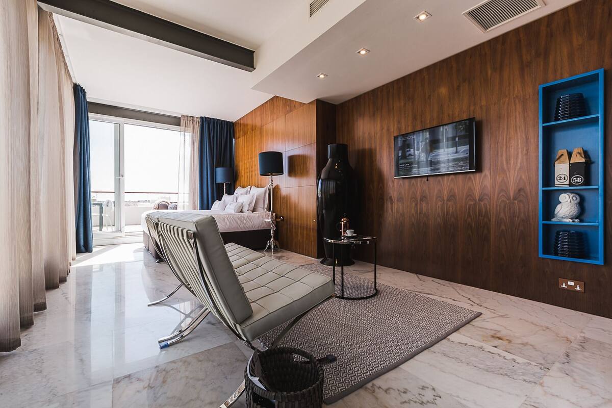 Una habitación de hotel moderna con una cama, una zona de estar, un ventanal grande y un televisor instalado en la pared.