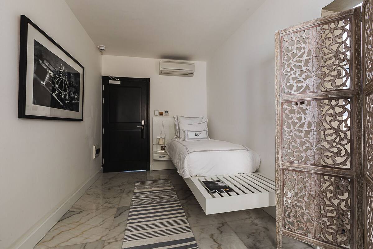 Habitación de hotel estrecha con una cama, una mesita de noche, un cuadro en la pared, un equipo de aire acondicionado y un biombo decorativo.