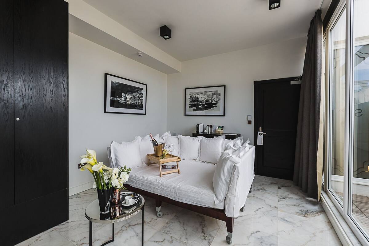 Una sala de estar moderna con un sofá blanco, una mesita con una botella de champán y un jarrón de flores. Hay dos fotografías en blanco y negro en la pared.