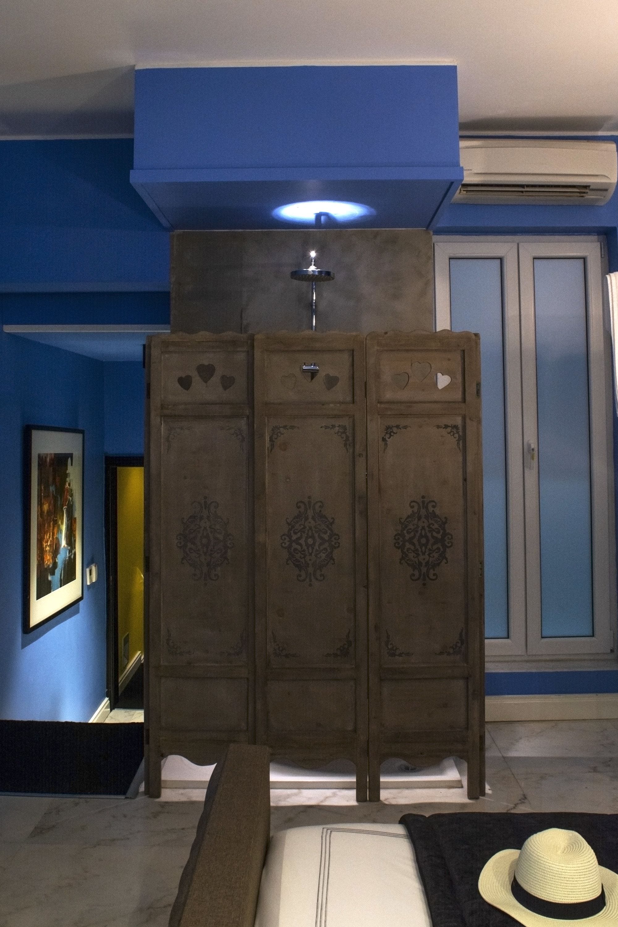 A room with a blue wall, a wooden cabinet with decorative panels, a bed with a white mattress and a brown blanket, and a hat on the bed.