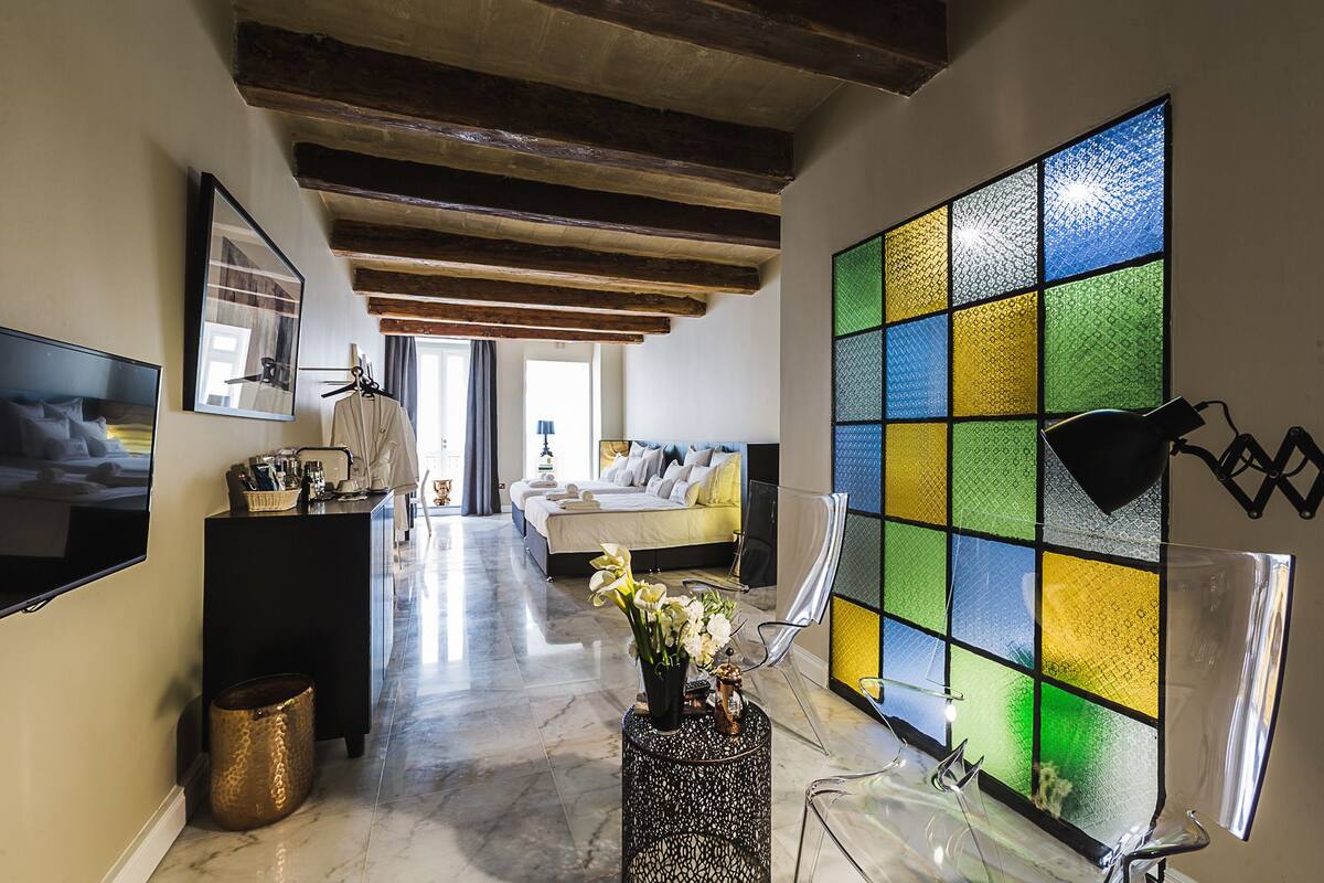 Habitación de hotel con cama, escritorio, televisor, un amplio ventanal con vitrales y un pasillo que conduce a otra habitación.