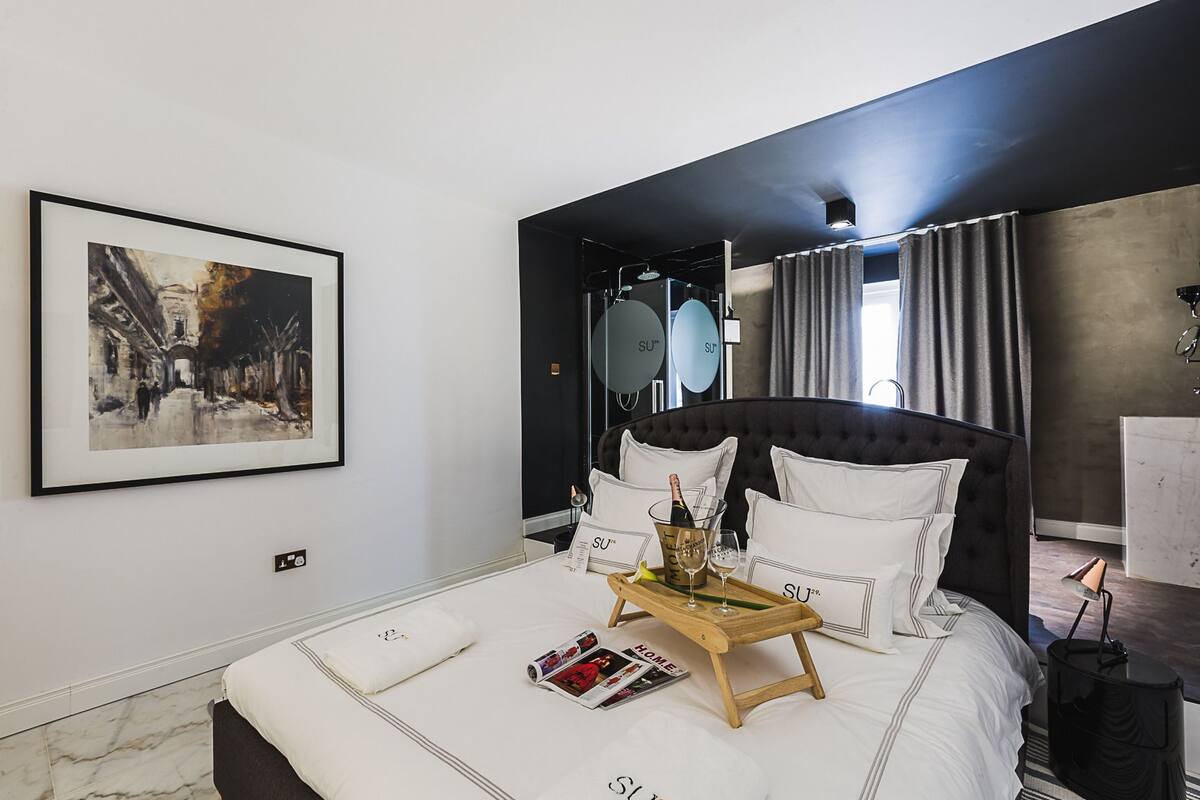 Una habitación de hotel moderna con una cama grande, un cuadro enmarcado con una vista urbana y un minibar, además de vistas a la ciudad desde la ventana.