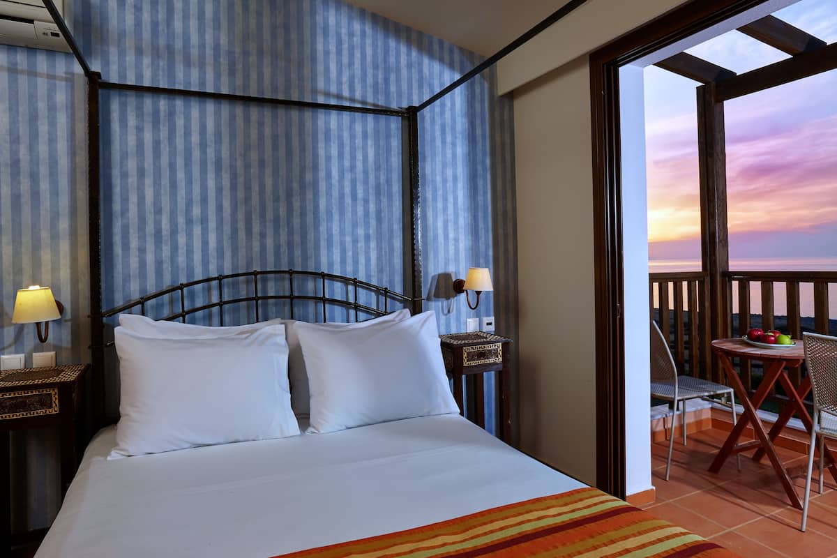 Een hotelkamer met een bed, een nachtkastje met een lamp, een tafeltje met een fruitschaal en een balkon met uitzicht op de zonsondergang.