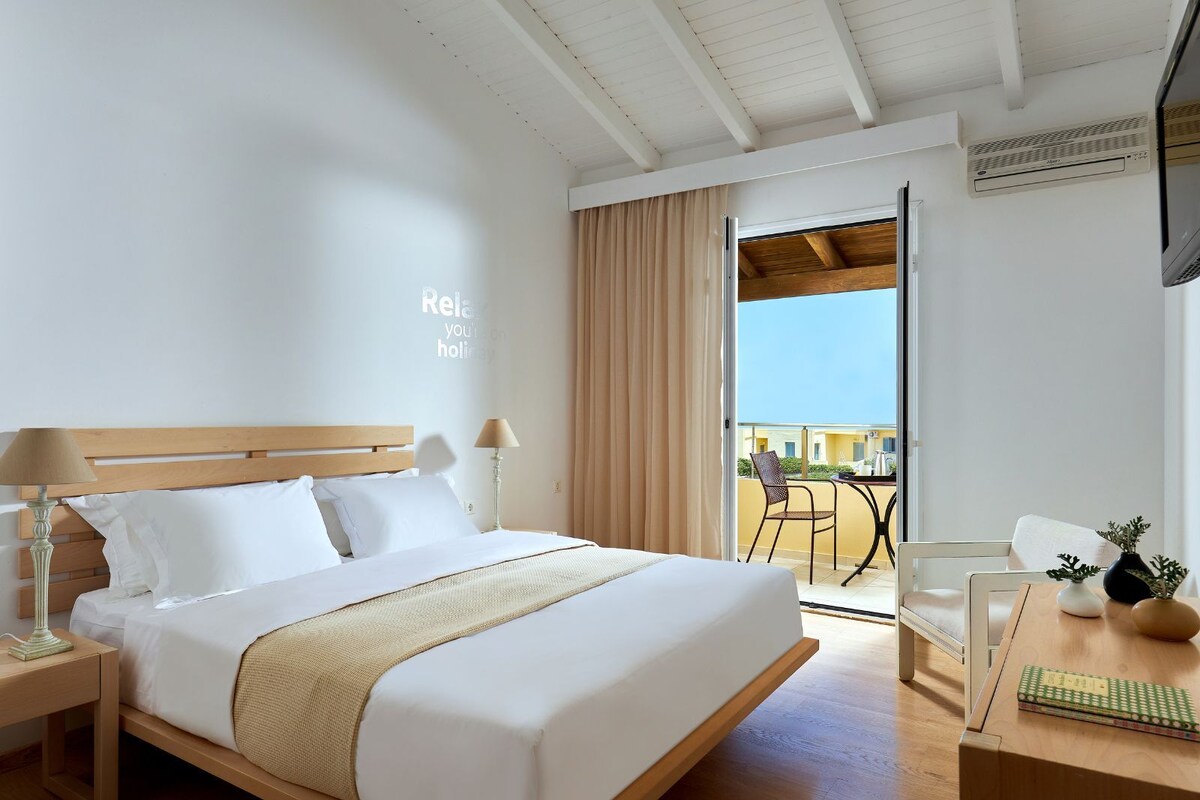 Hotelkamer met een bed, bedlampen, een bureau met een stoel, een klein tafeltje met een vaas en een boek, en een balkon met een stoel en tafeltje.