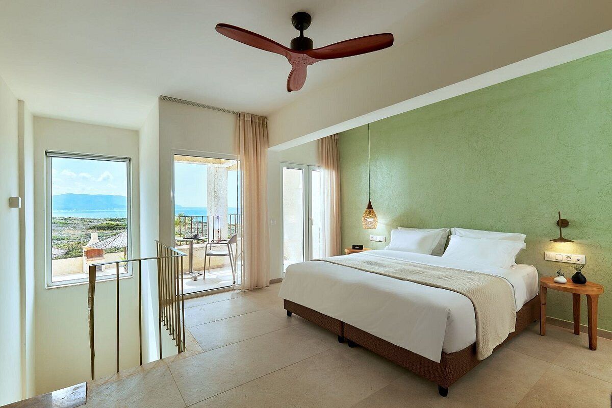 A bedroom with a bed, a ceiling fan, a nightstand, a lamp, and a view of the ocean.