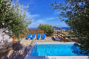 Un área de piscina con sillas reclinables azules, una pérgola de madera y vistas al mar.