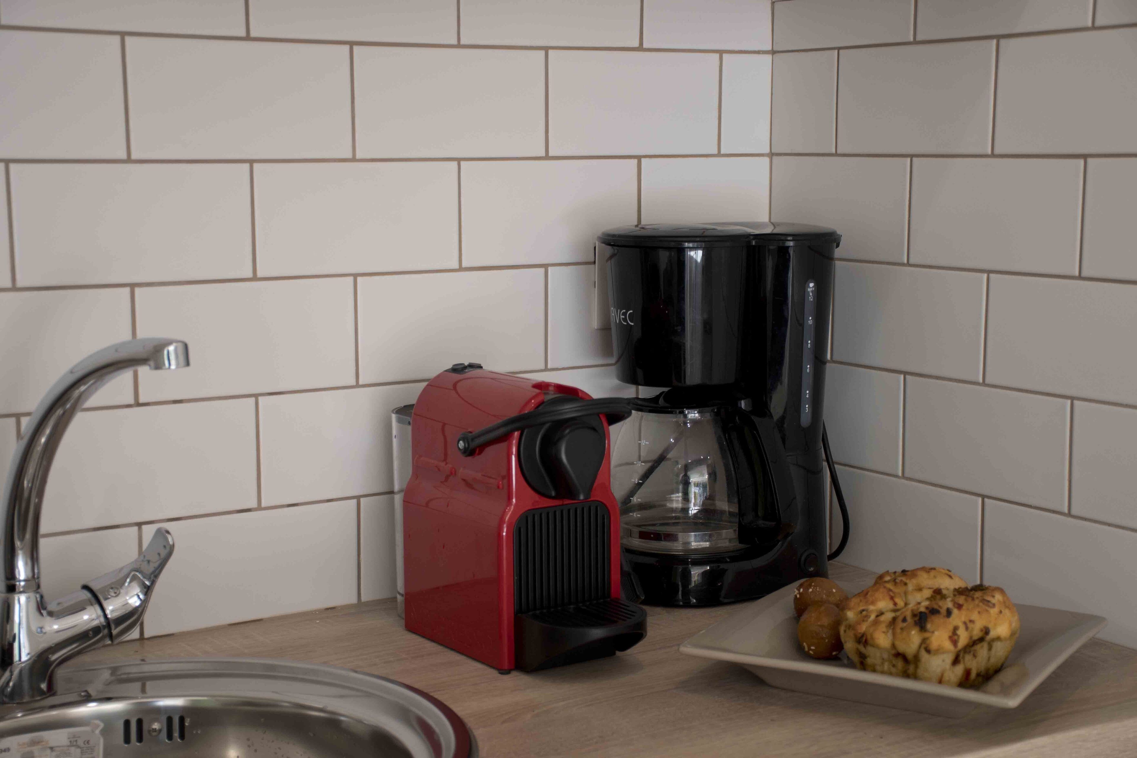 Een keuken met een spoelbak, een rode koffiemachine, een zwarte koffiepot en een bord met twee gebakjes.