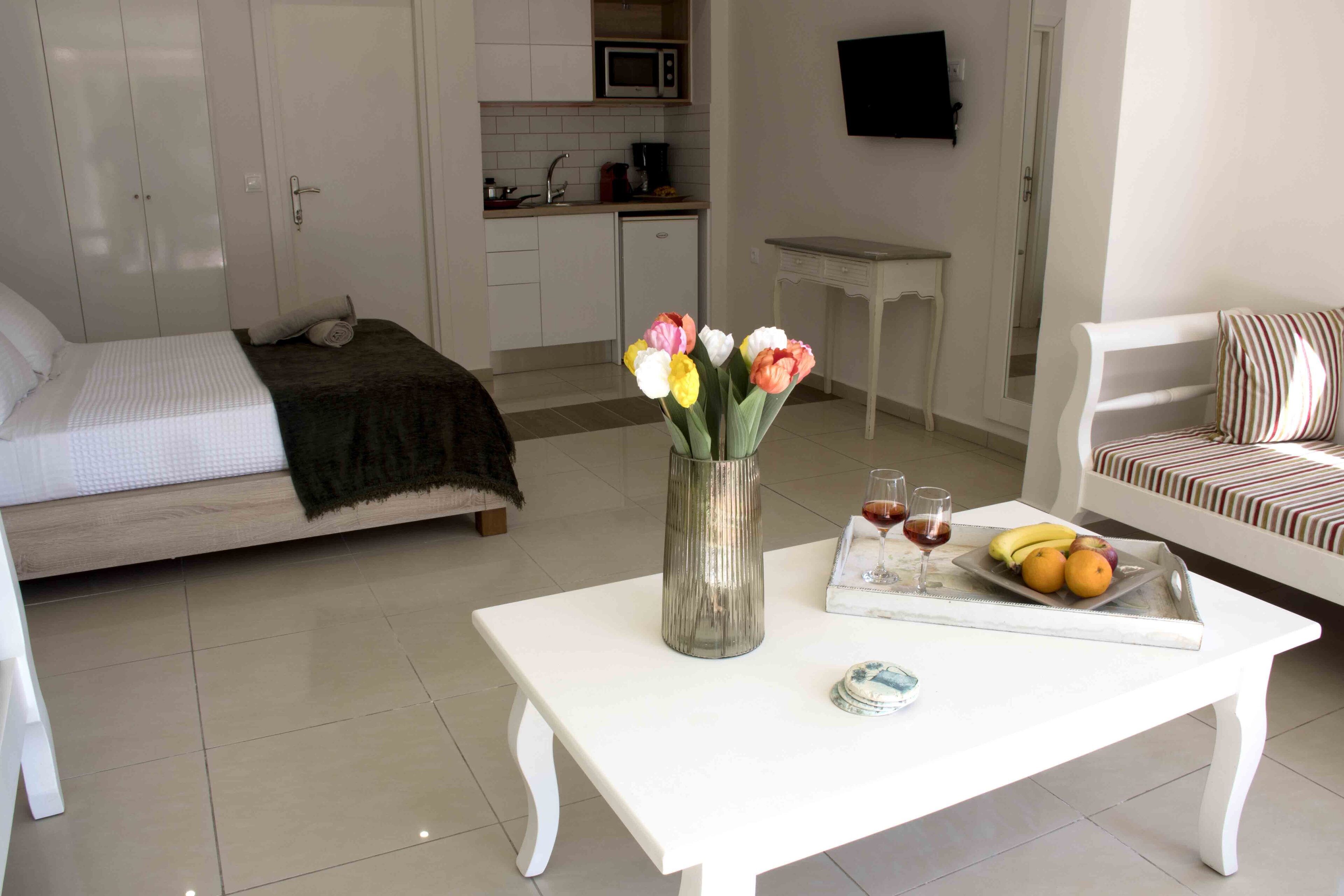 A bedroom with a bed, a bedside table with a vase of flowers, a tray with fruits and glasses, and a view of the kitchen and living area.