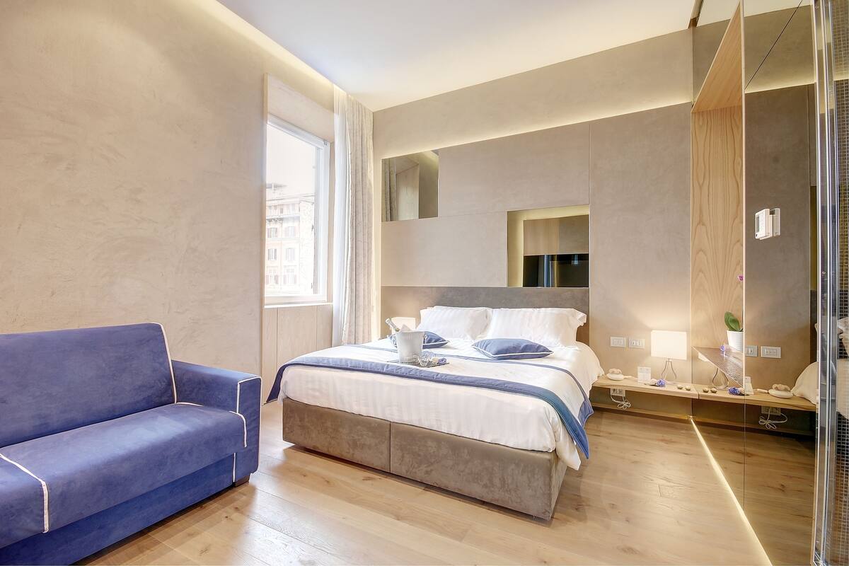 A modern hotel room with a large bed, a blue sofa, a bedside table with a lamp, and a large window.