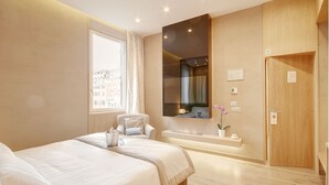 Una habitación de hotel moderna con una cama grande, una zona de estar con una silla, una mesita, un ventanal con vistas a edificios, un espejo y una puerta de madera.