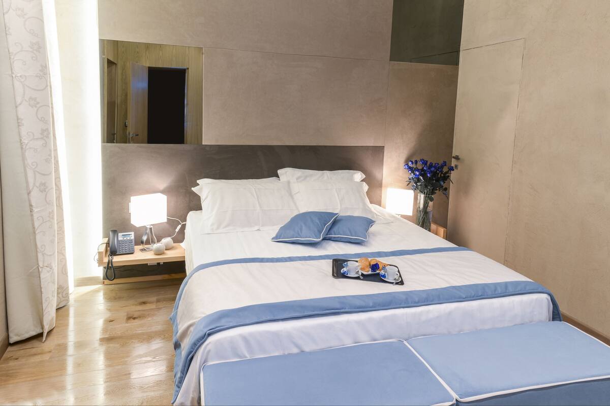 A neatly arranged hotel room with a large bed, bedside lamps, a bedside table with a phone, and a tray with breakfast items.