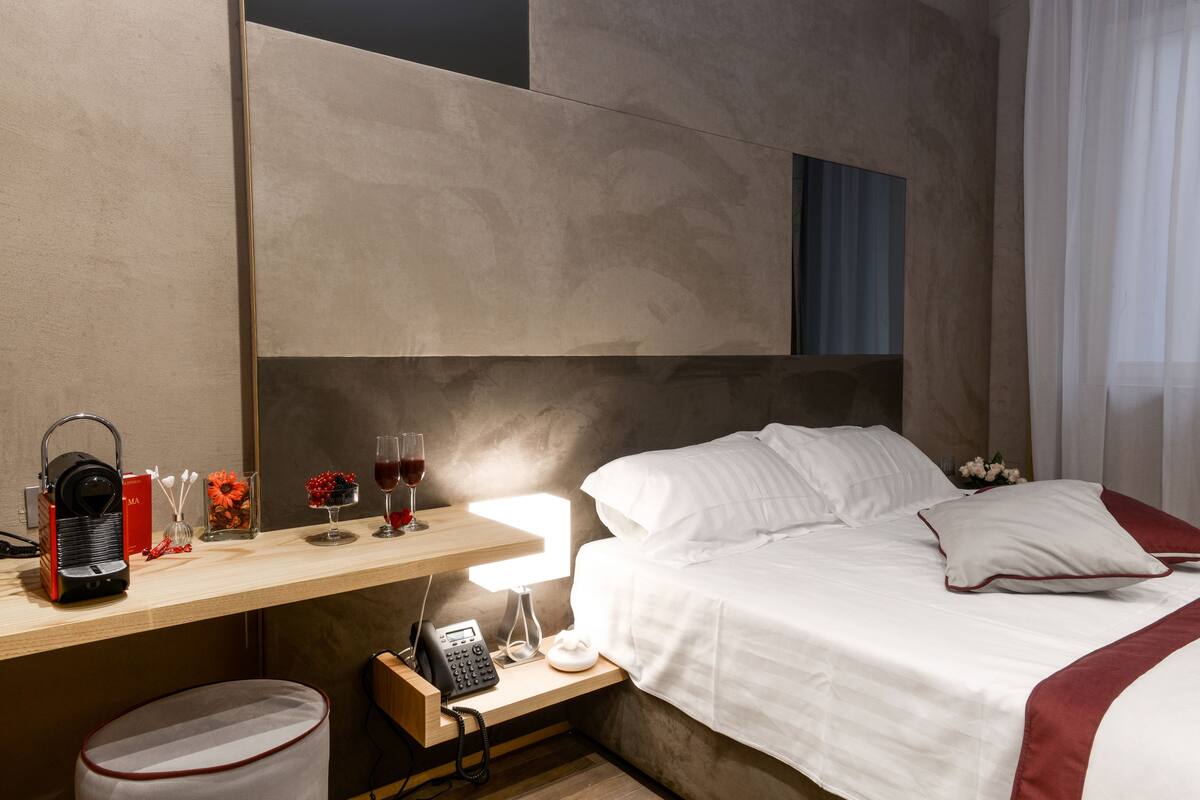 A hotel room with a bed, bedside table, lamp, and a small desk area with a phone, a red box, and a glass container with flowers.