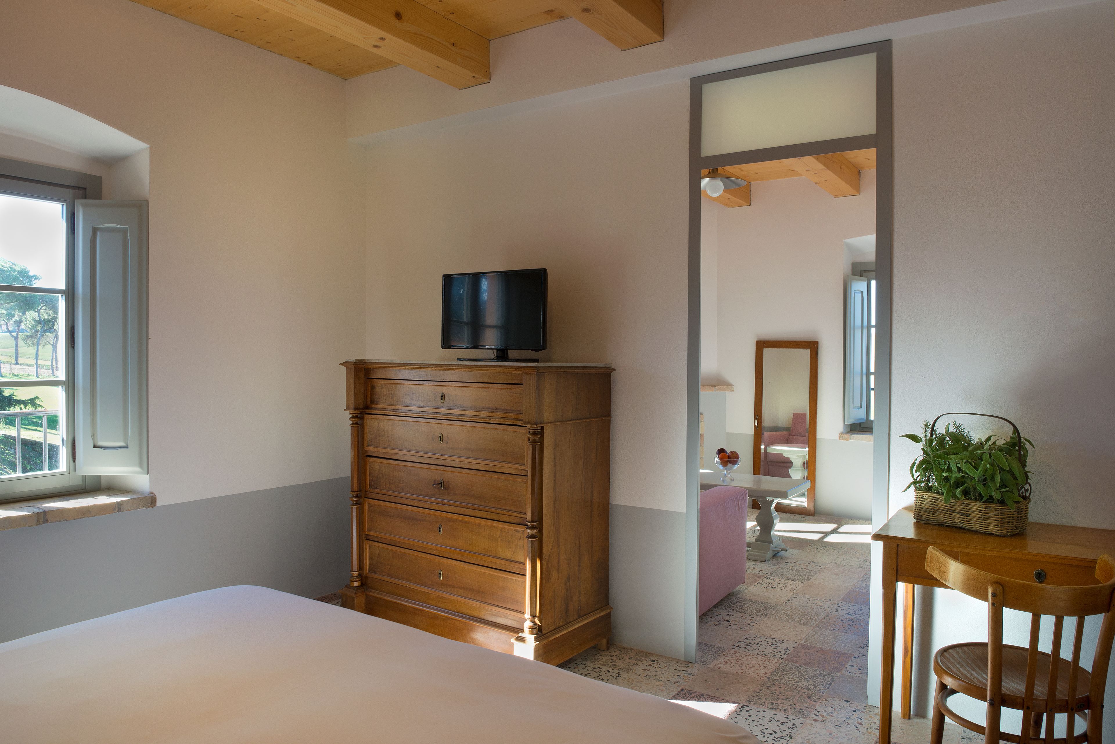 Un dormitorio con cama, cómoda de madera, televisor encima, ventana, espejo y una mesita con una planta.