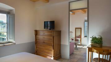Un dormitorio con cama, cómoda de madera, televisor encima, ventana, espejo y una mesita con una planta.