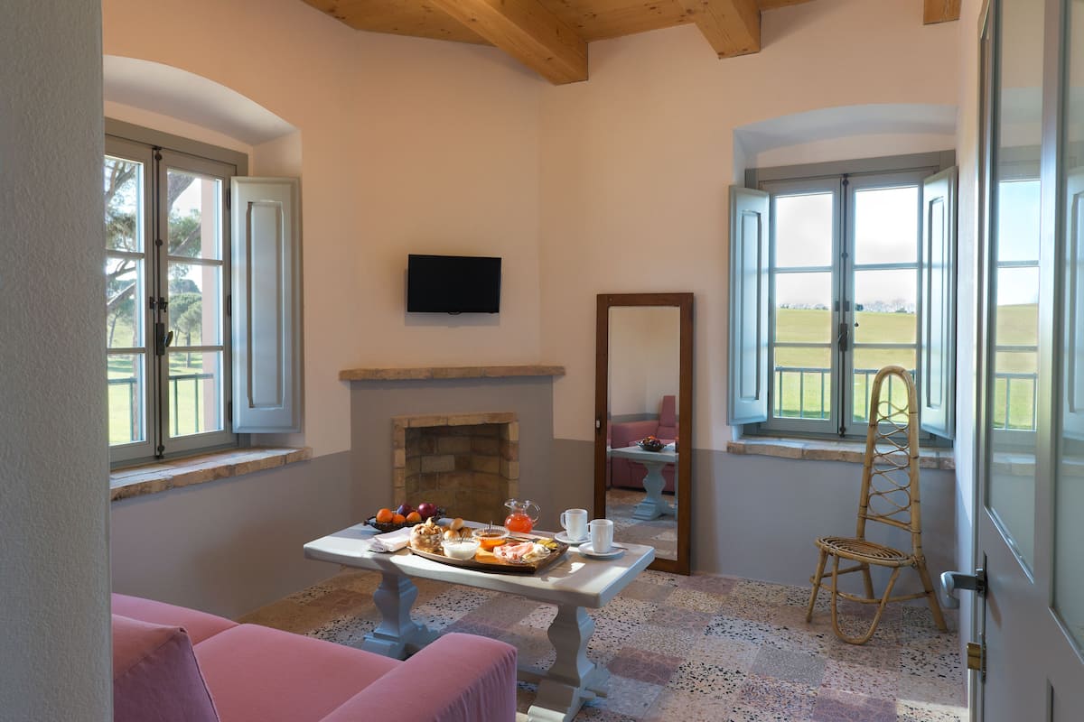 A room with a breakfast table set with food, a television on the wall, and a view of the outdoors through large windows.