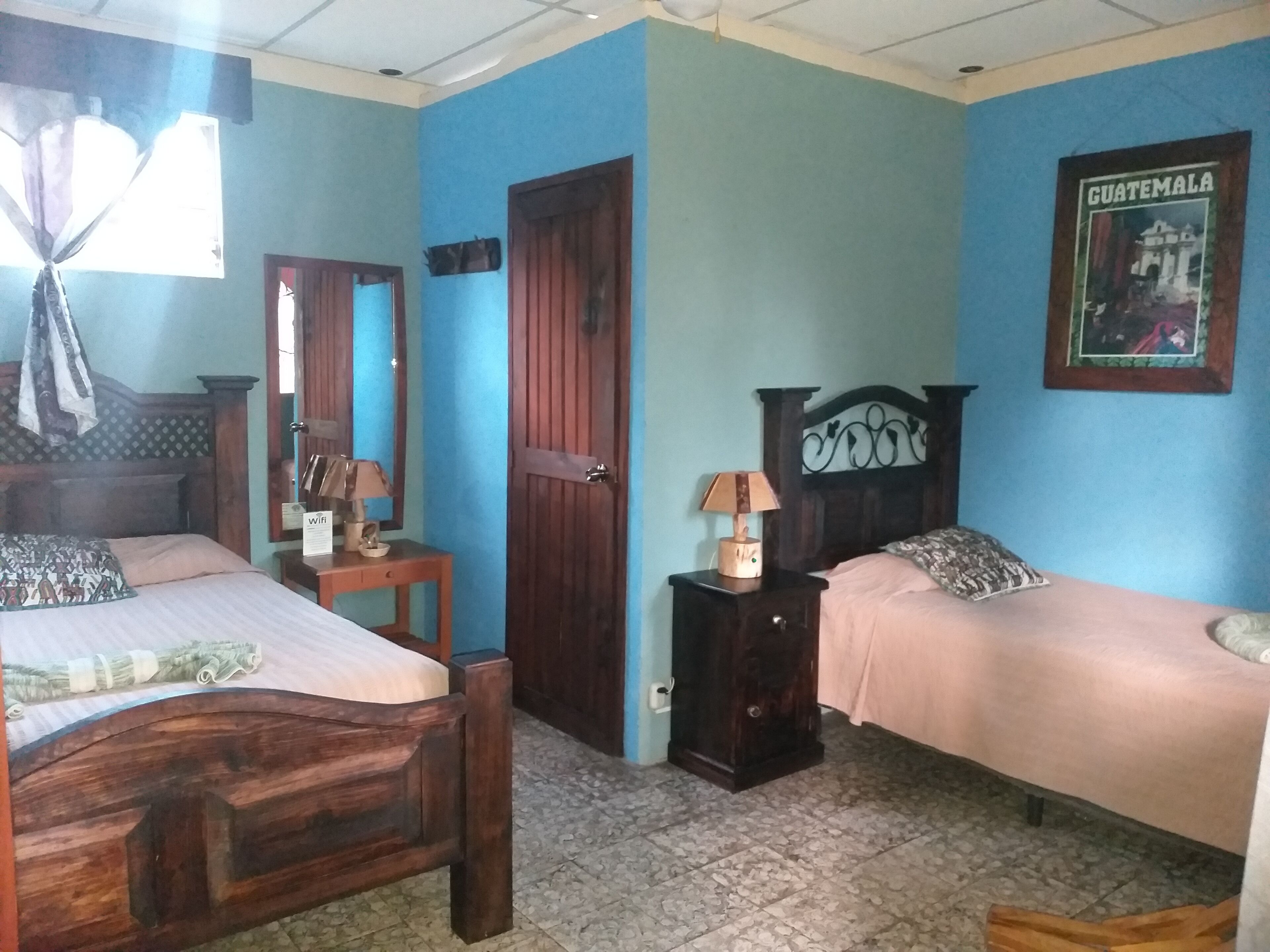 Una stanza con due letti di legno, un comodino con una lampada, una porta e un quadro appeso al muro raffigurante la Guatemala.