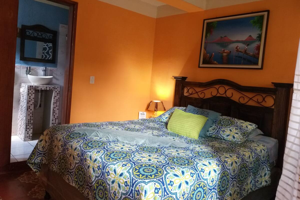 Une chambre à coucher comprenant un lit, une table de chevet, un tableau, et une salle de bain avec un lavabo et un miroir.