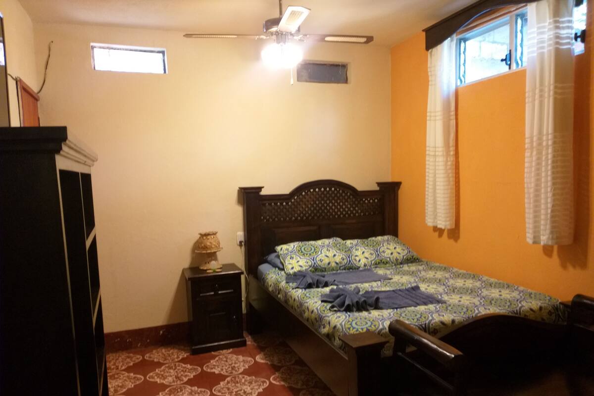 Une chambre à coucher comprenant un lit, une table de chevet, une commode, une fenêtre avec des rideaux et un ventilateur de plafond.