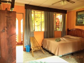 A room with a bed, a wooden chair, a window with curtains, a ceiling fan, and a painting on the wall. - Hospedaje El Viajero (Panajachel)