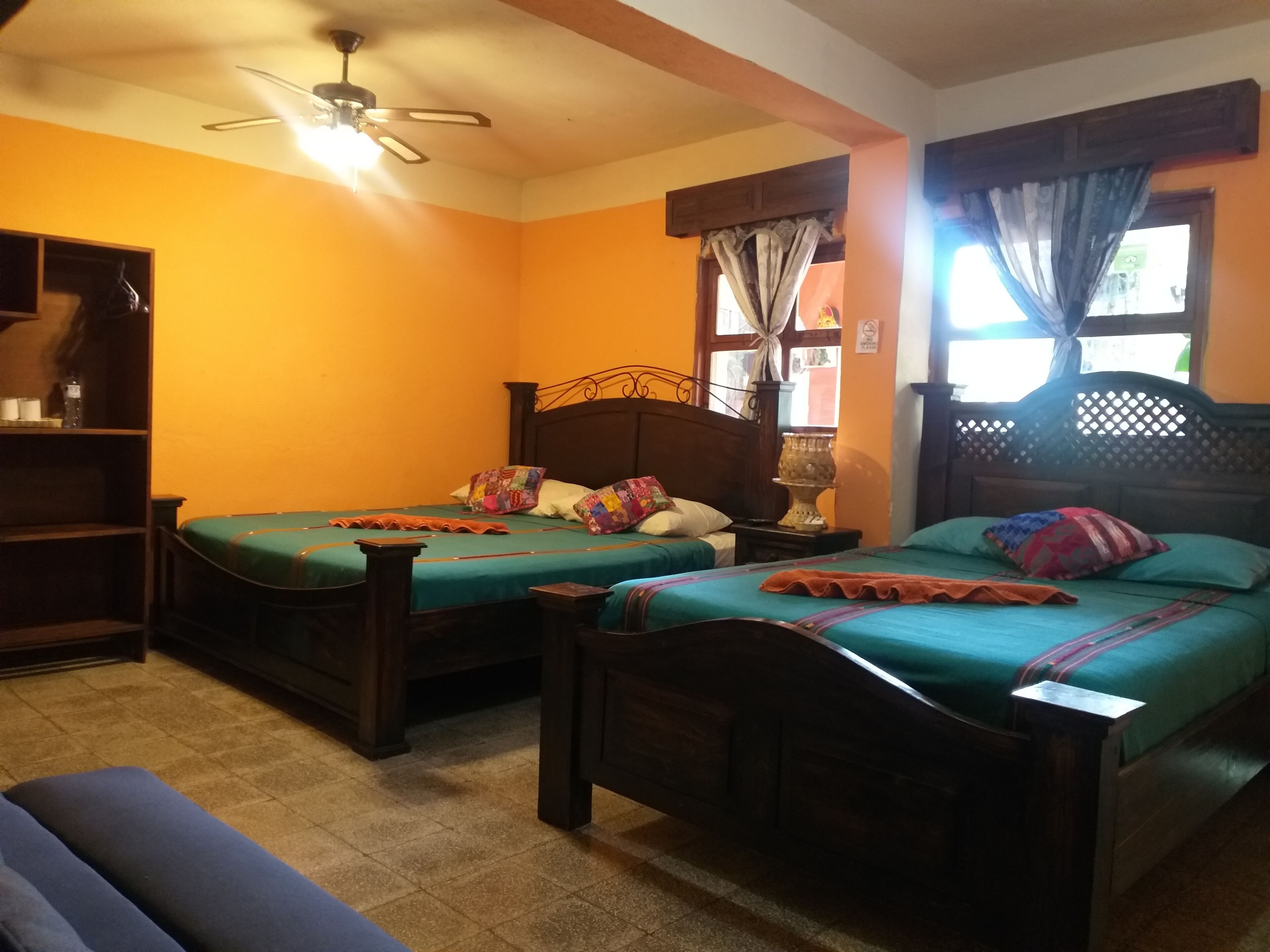 A room with two wooden beds, a ceiling fan, a window with curtains, and a shelf with items.