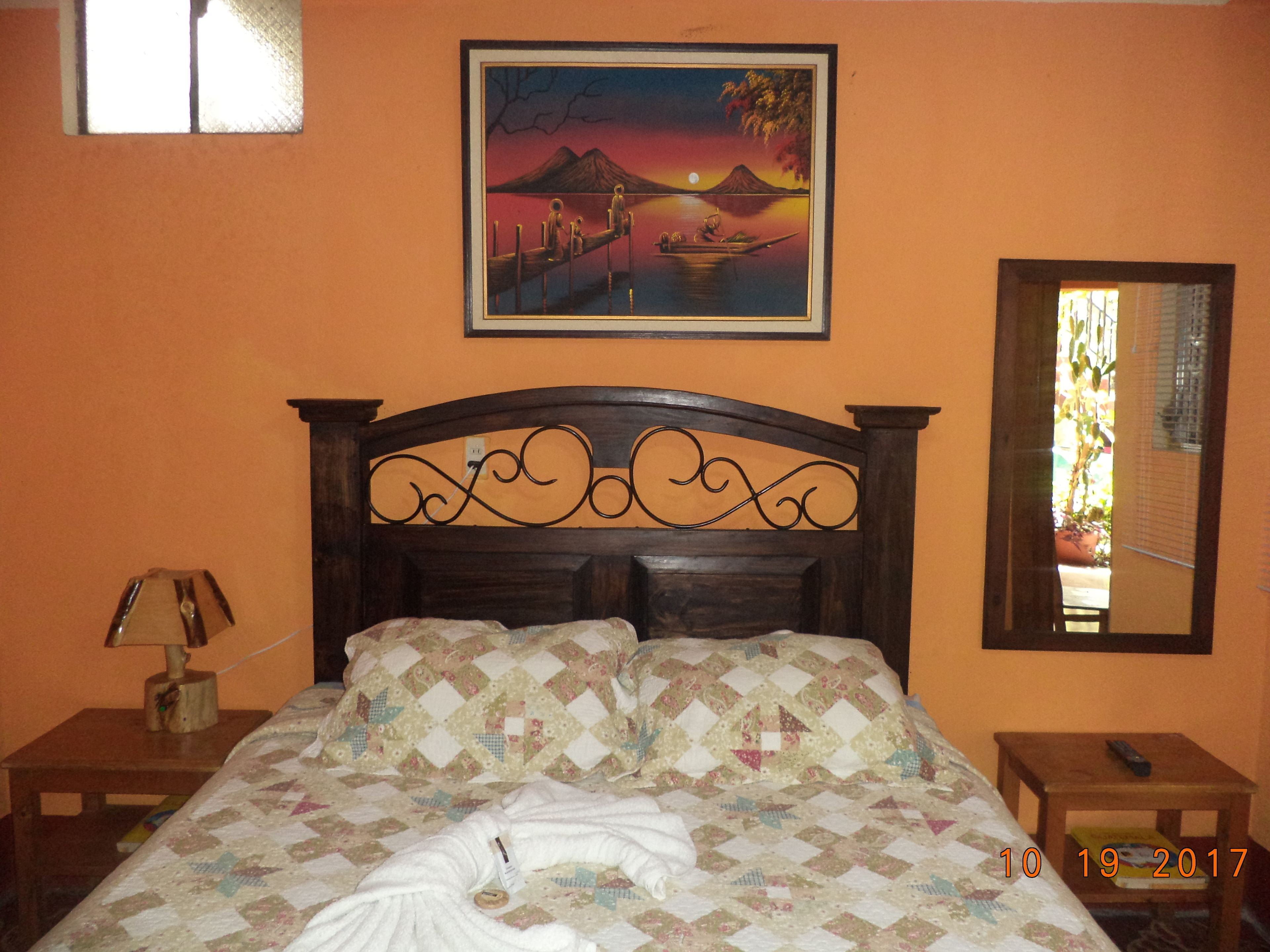 Un letto rifatto con un copriletto, una lampada sul comodino, un quadro incorniciato con una veduta al tramonto e uno specchio appeso al muro.