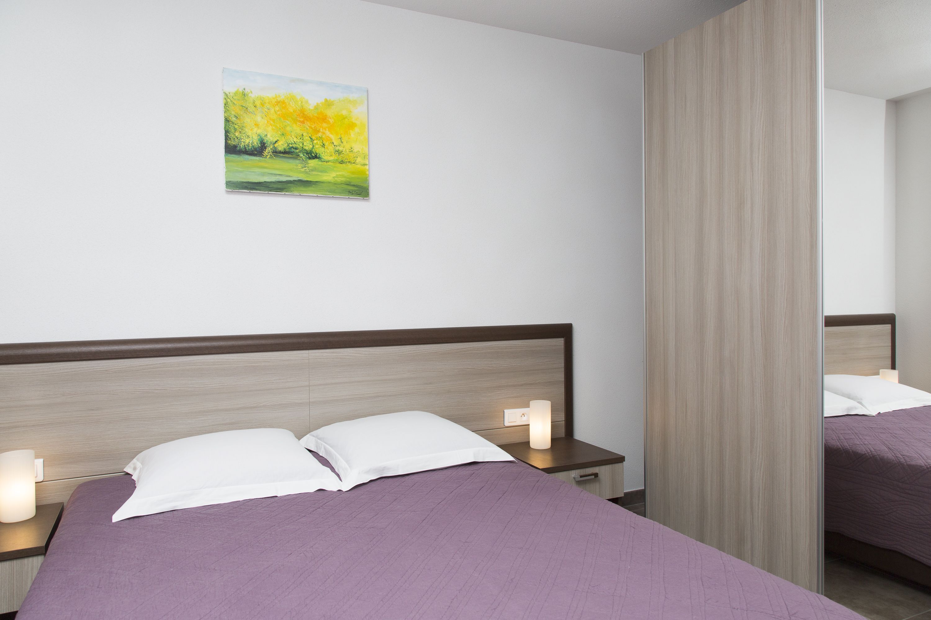 Ein ordentlich bezogenes Bett mit einem violetten Überwurf, zwei weißen Kissen und einem Nachttisch mit einer Lampe.