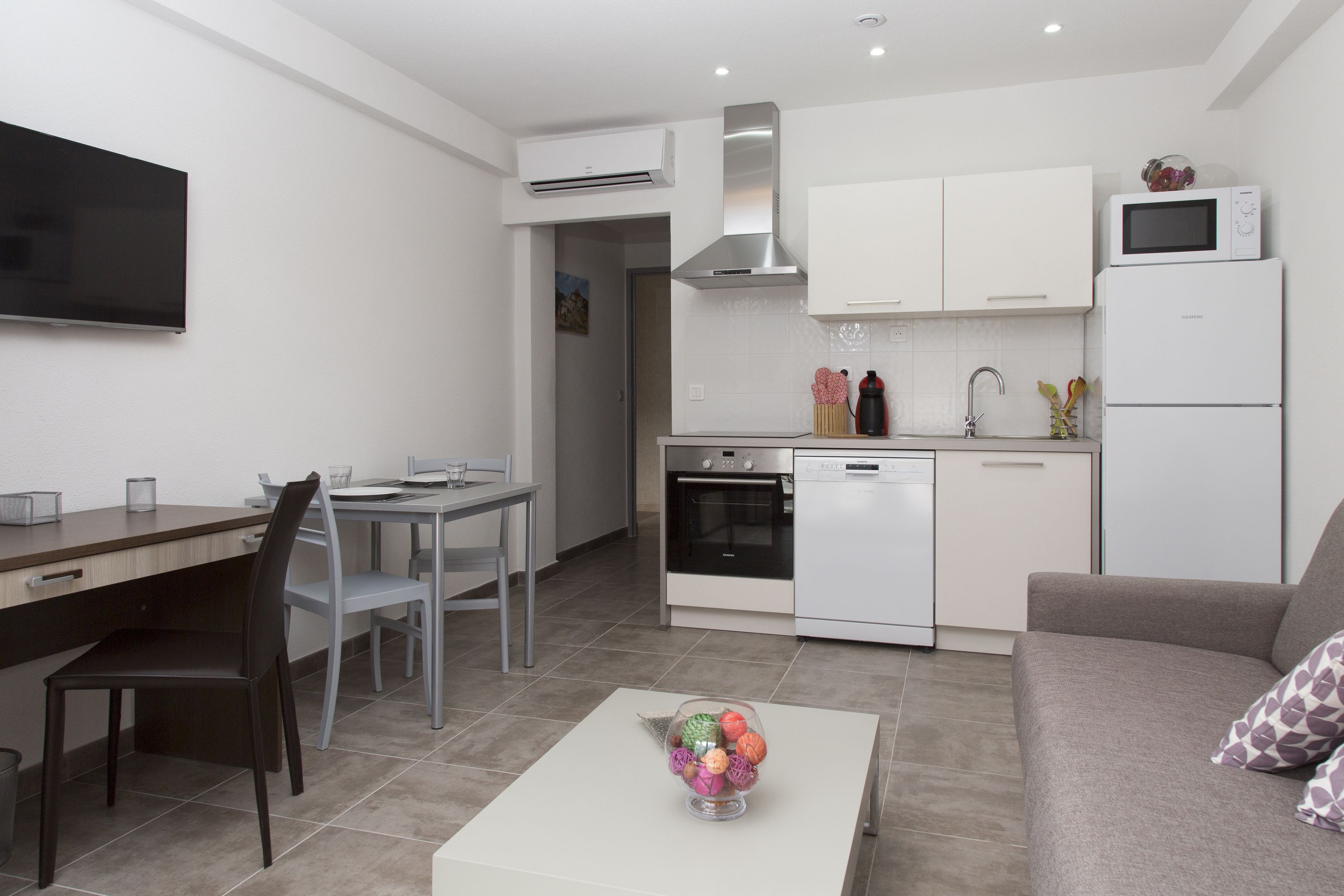 A modern kitchen with white cabinets, a microwave, and a dishwasher. There is a dining area with a table and chairs, and a living area with a sofa and a coffee table.