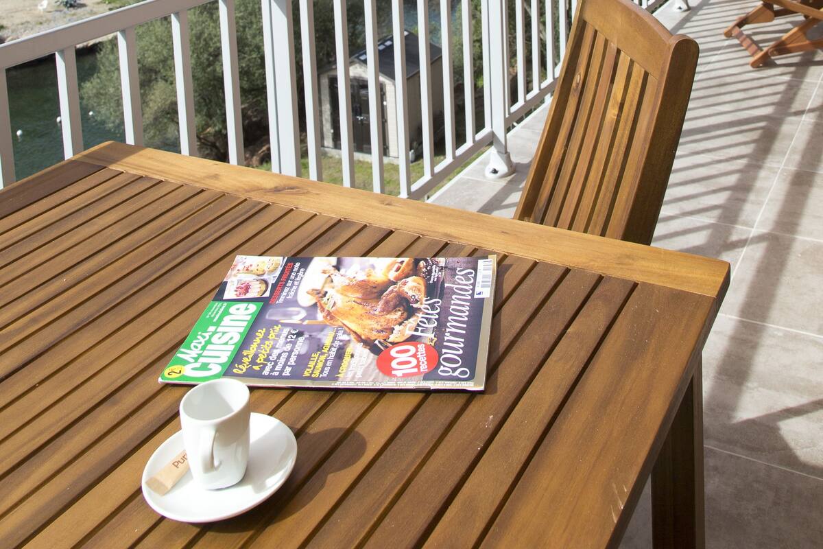 Een houten tafel met een menukaart en een kopje op een balkon met uitzicht op een bergachtig landschap en een strand.