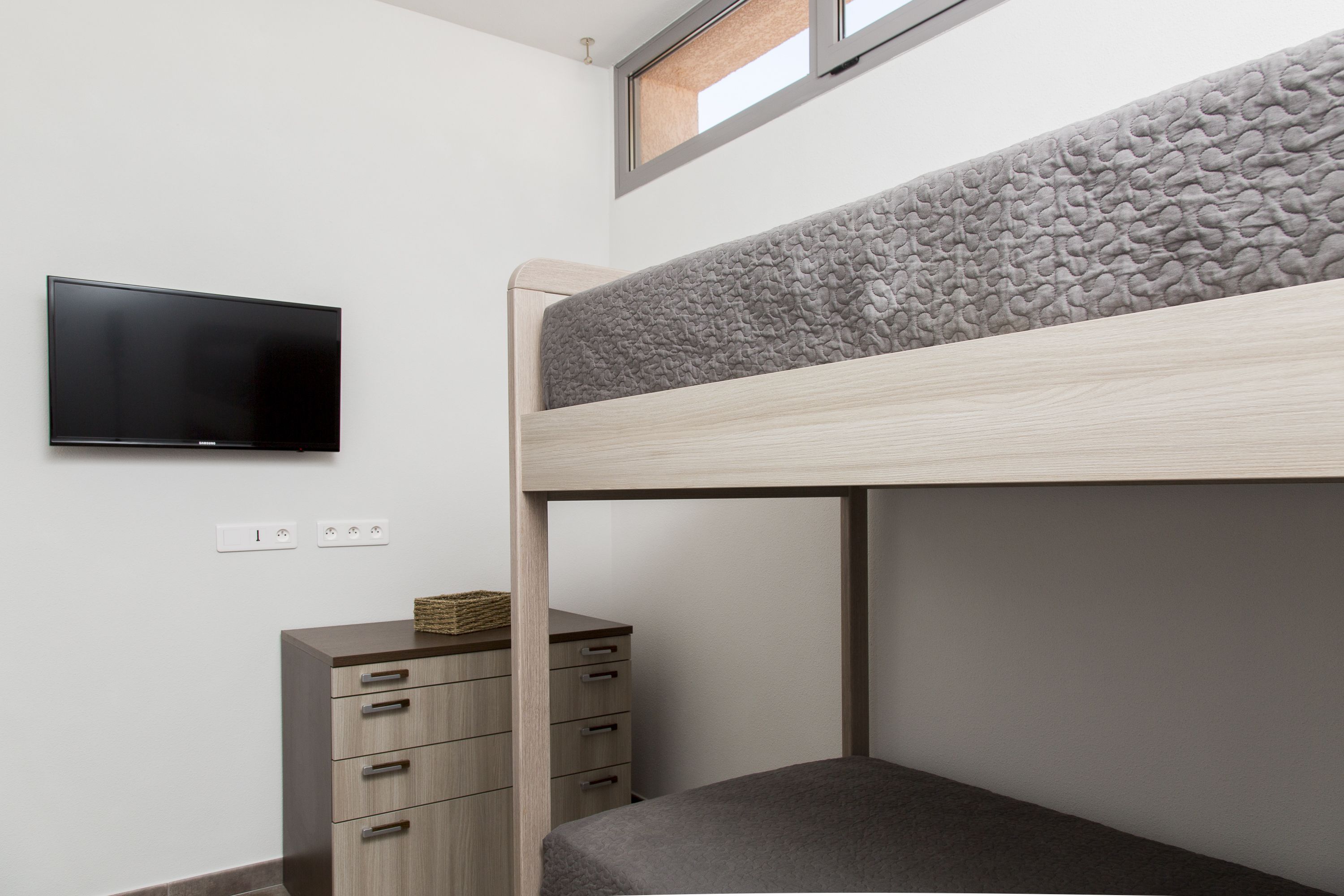 A bunk bed with a television mounted on the wall, a wooden dresser, and a window above the bed.