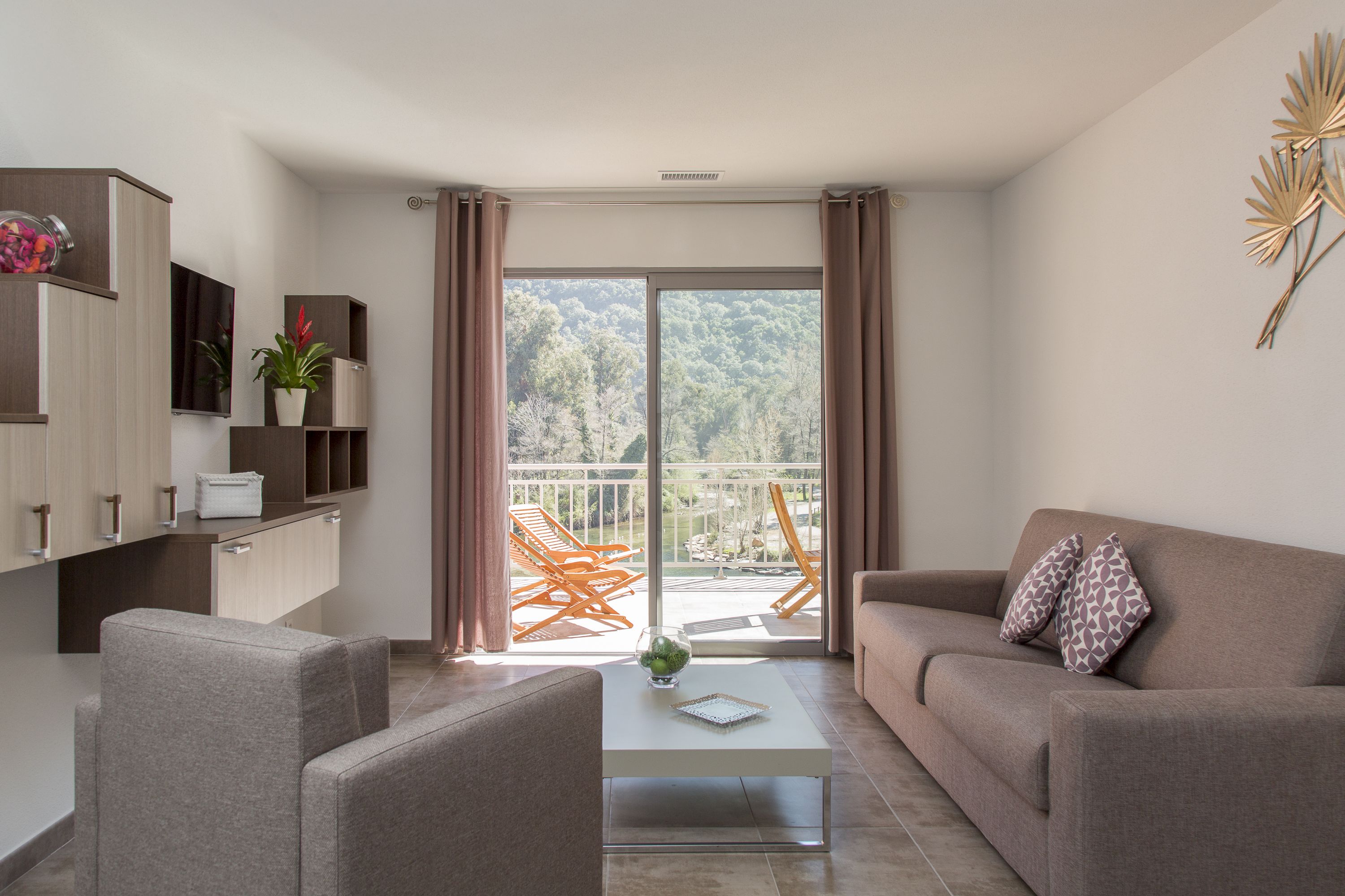Un soggiorno moderno con divano, poltrone, tavolino e TV su un mobile a parete. Un balcone con vista sugli alberi e una sedia all'esterno.