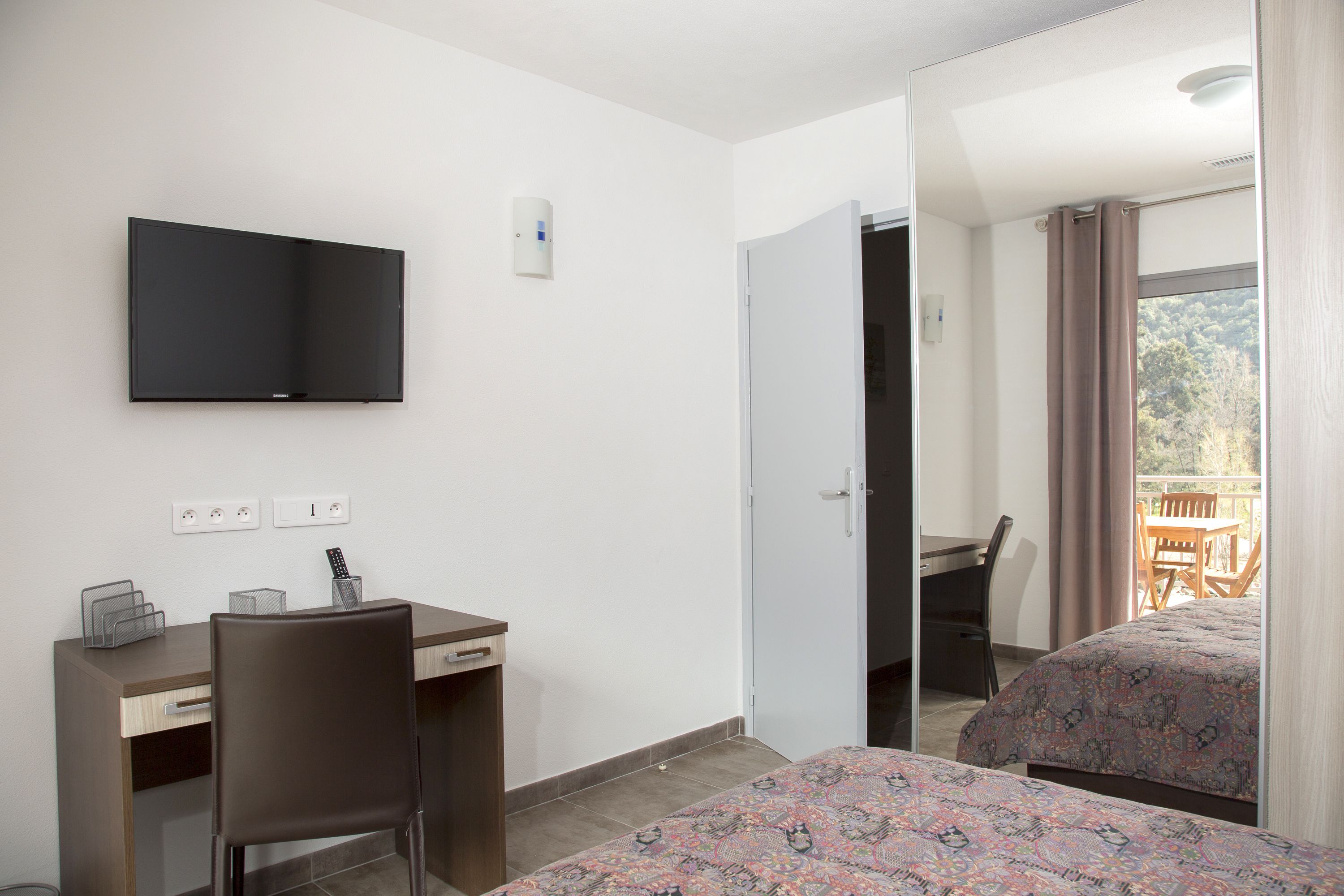 Une chambre d’hôtel comprenant un lit, un bureau avec une chaise, un téléviseur fixé au mur et un balcon avec une table et des chaises.