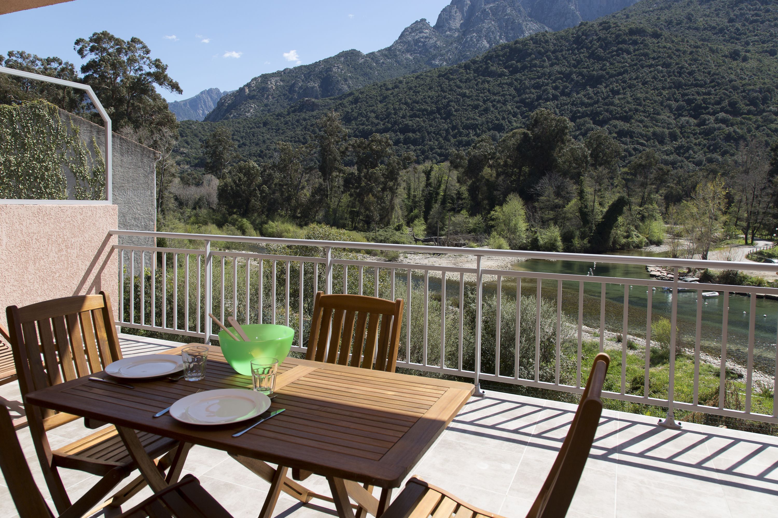 Un balcón con una mesa dispuesta para dos, con vista a un río y montañas.