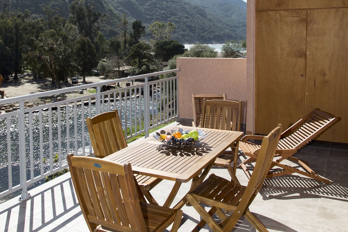 Een balkon met een houten tafel en stoelen, met uitzicht op een bergachtig landschap en een wateroppervlak.