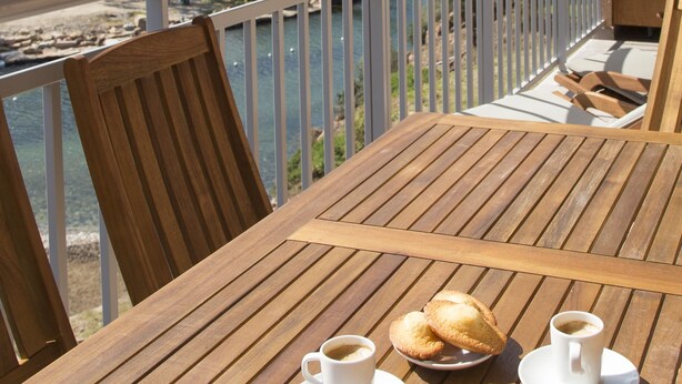A wooden table with two cups of coffee and two pastries on a balcony overlooking a lake and mountains.