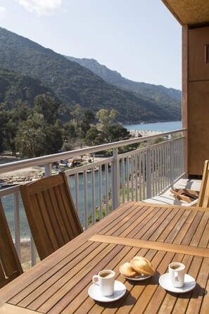 Una mesa de madera con dos tazas de café y dos pasteles en un balcón con vista a un lago y montañas.