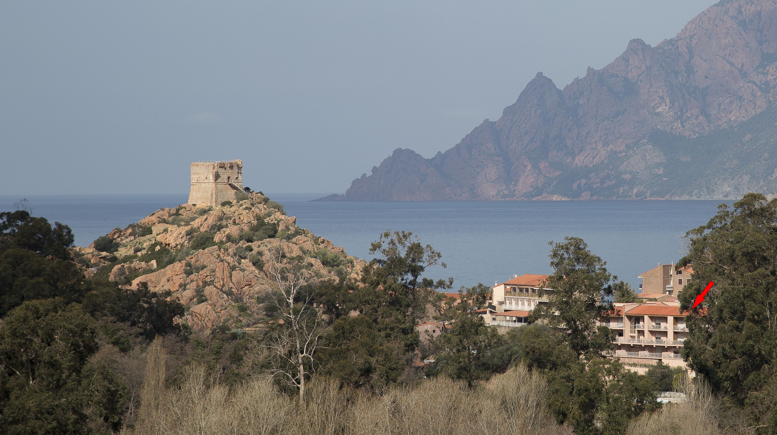 Un paysage côtier avec une tour de pierre historique située sur une colline, une mer en arrière-plan et un immeuble résidentiel sur lequel pointe une flèche rouge.
