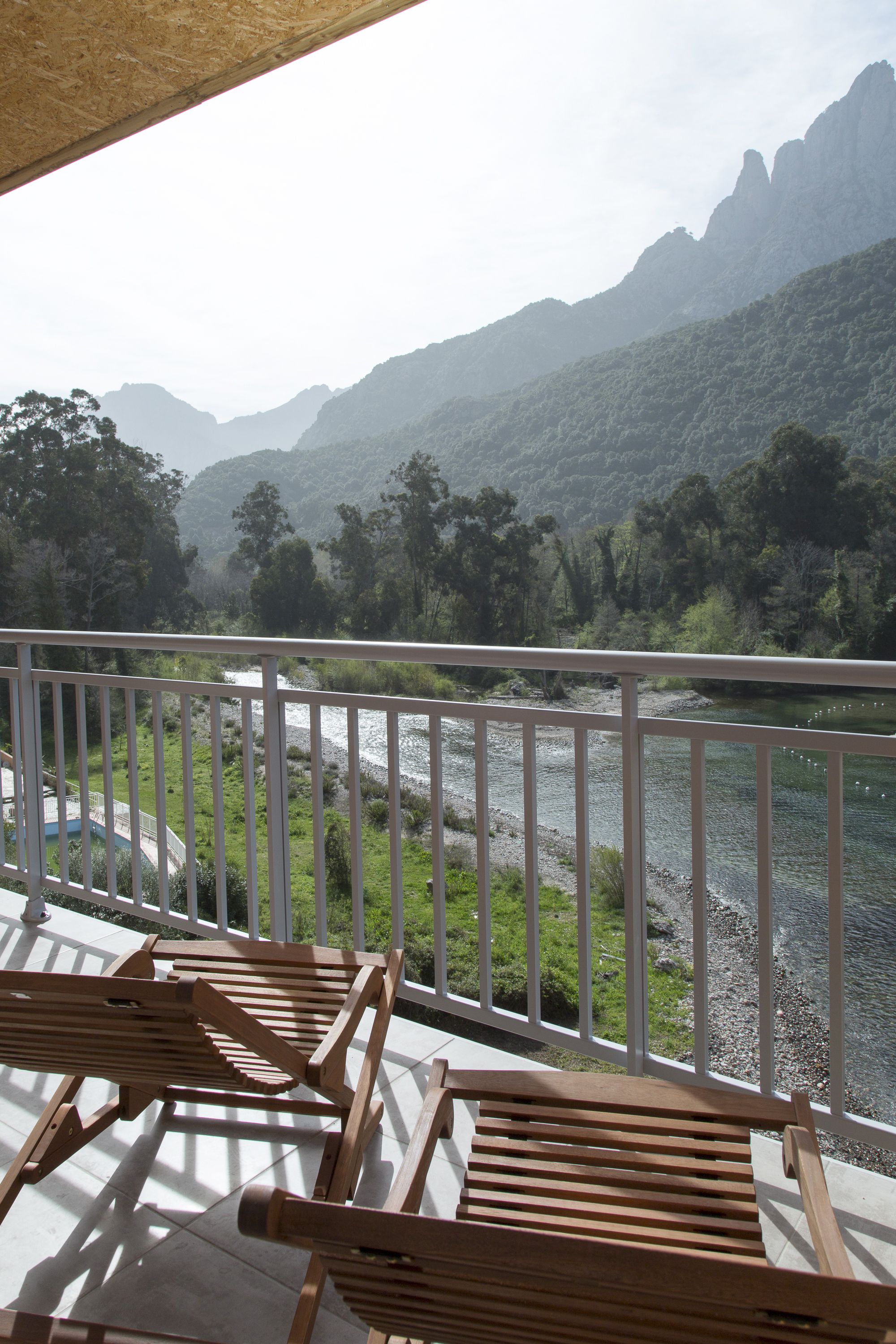 A balcony with wooden chairs overlooking a river and mountains.
