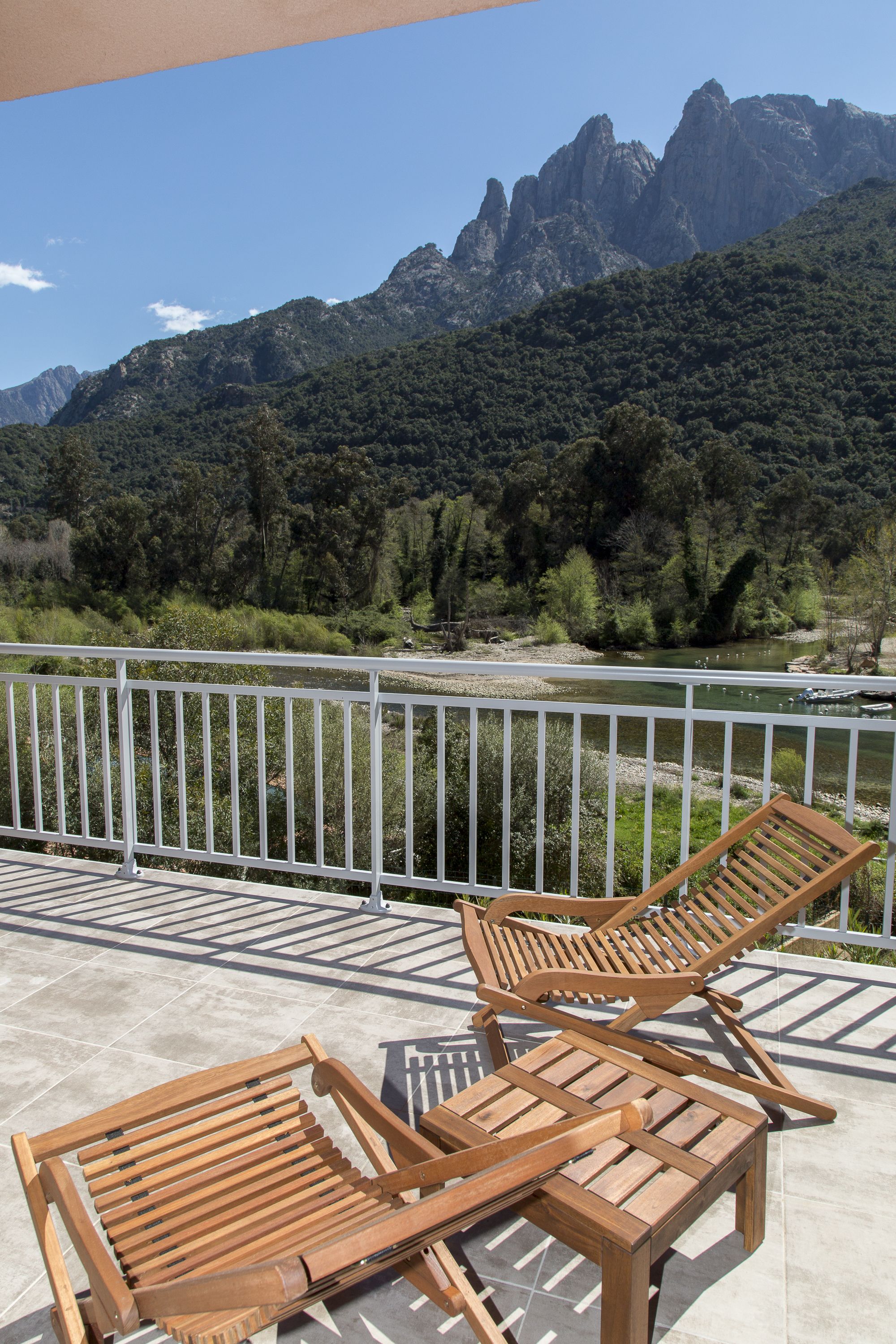 A balcony with two wooden lounge chairs overlooking a river and mountains.