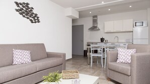 A modern living room with a grey sofa and armchair, a white coffee table with a plant, and a kitchen in the background.
