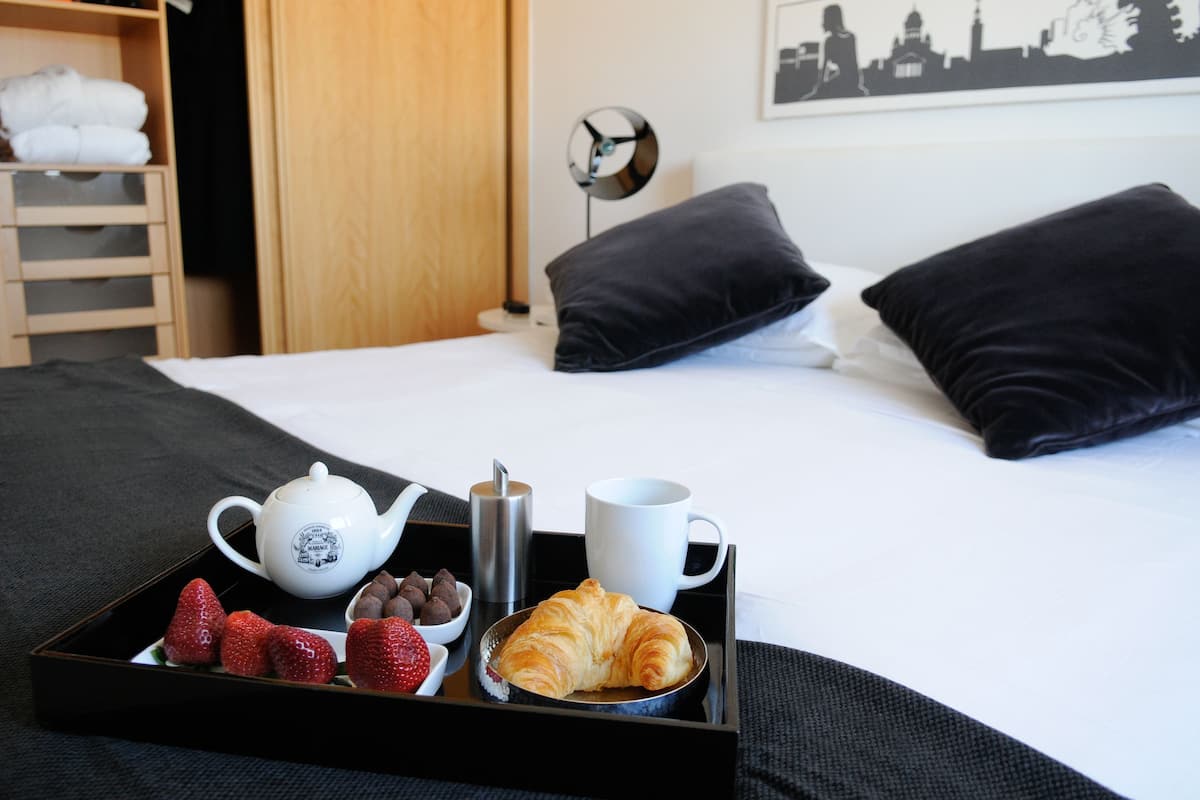 Una cama bien hecha con almohadas negras, una bandeja con tetera, taza, fresas y un croissant.