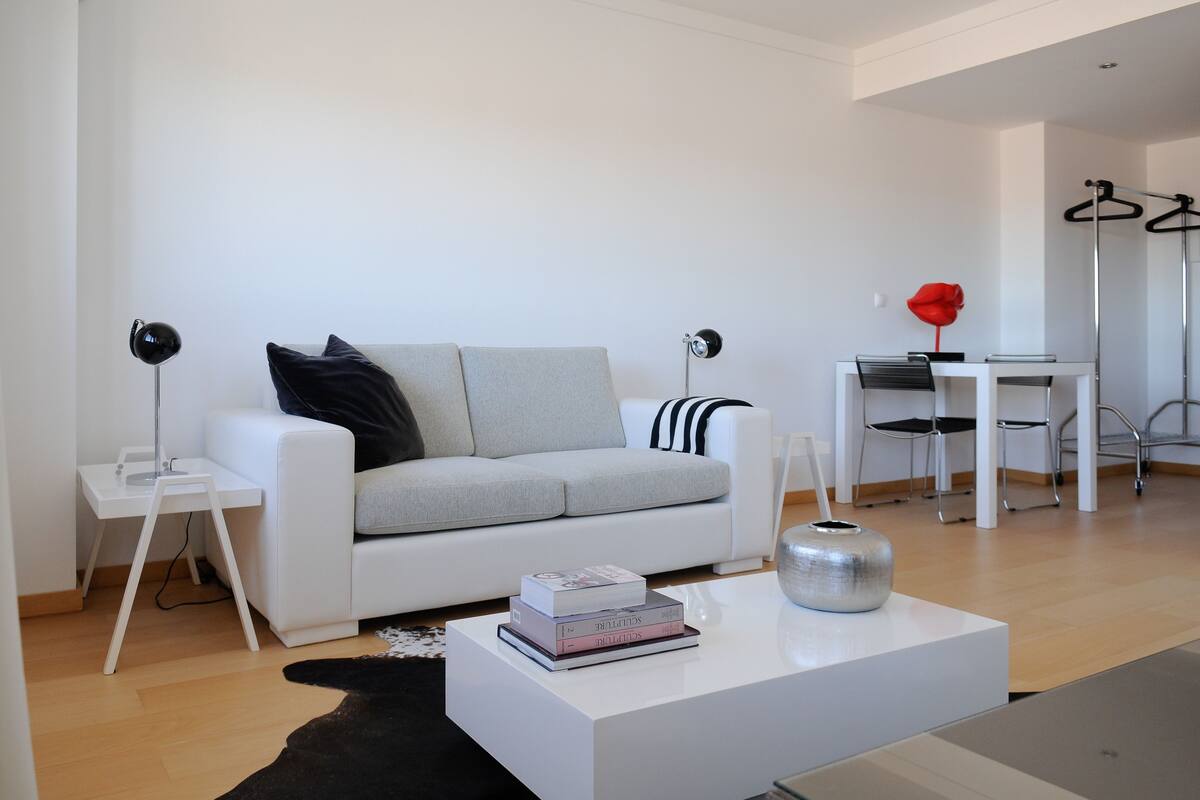 Un salón moderno con un sofá blanco, una mesa de centro con libros, un cojín a rayas negro y blanco, y un jarrón rojo.