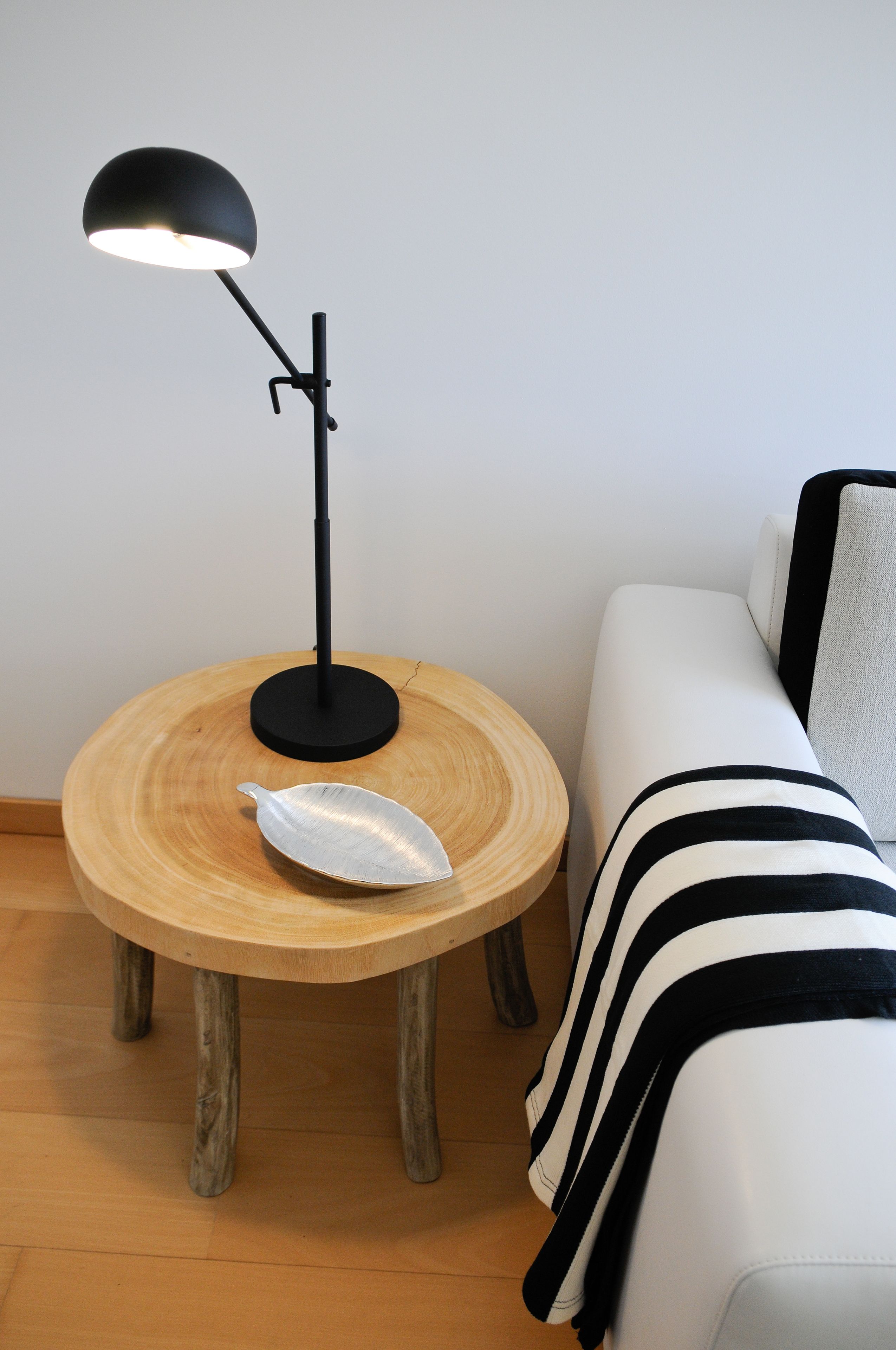 A round wooden table with a black lamp, a white cushion, and a striped throw blanket on a white sofa.