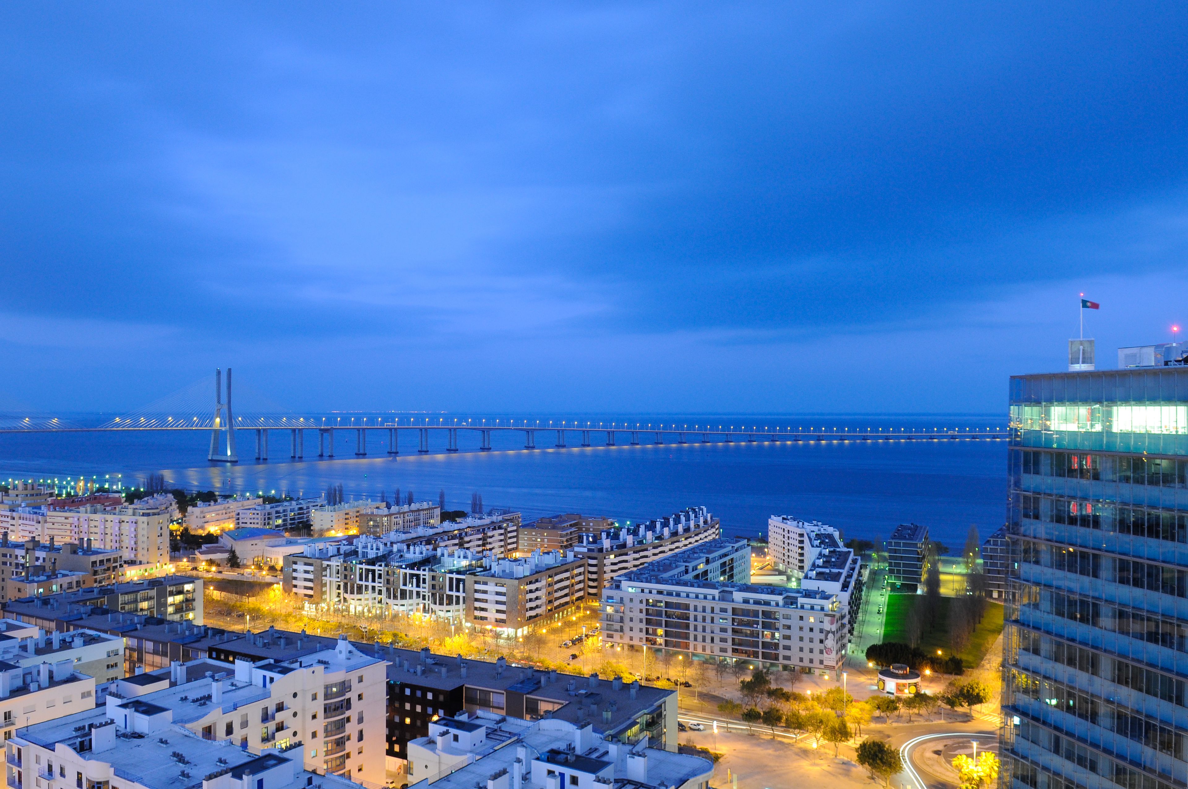 A cityscape at dusk with a prominent bridge in the distance, modern buildings, and a view of the sea.