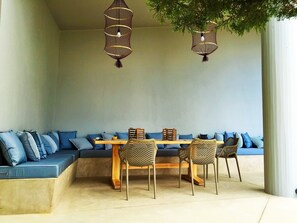 Un área de estar con una mesa y sillas de madera, un banco con cojines azules y lámparas colgantes.